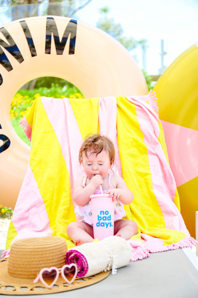 baby girl sipping through a straw in front of colorful towels and pool floats 