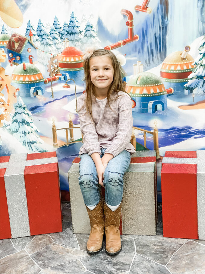 little girl posing on present box seats in santa's workshop 